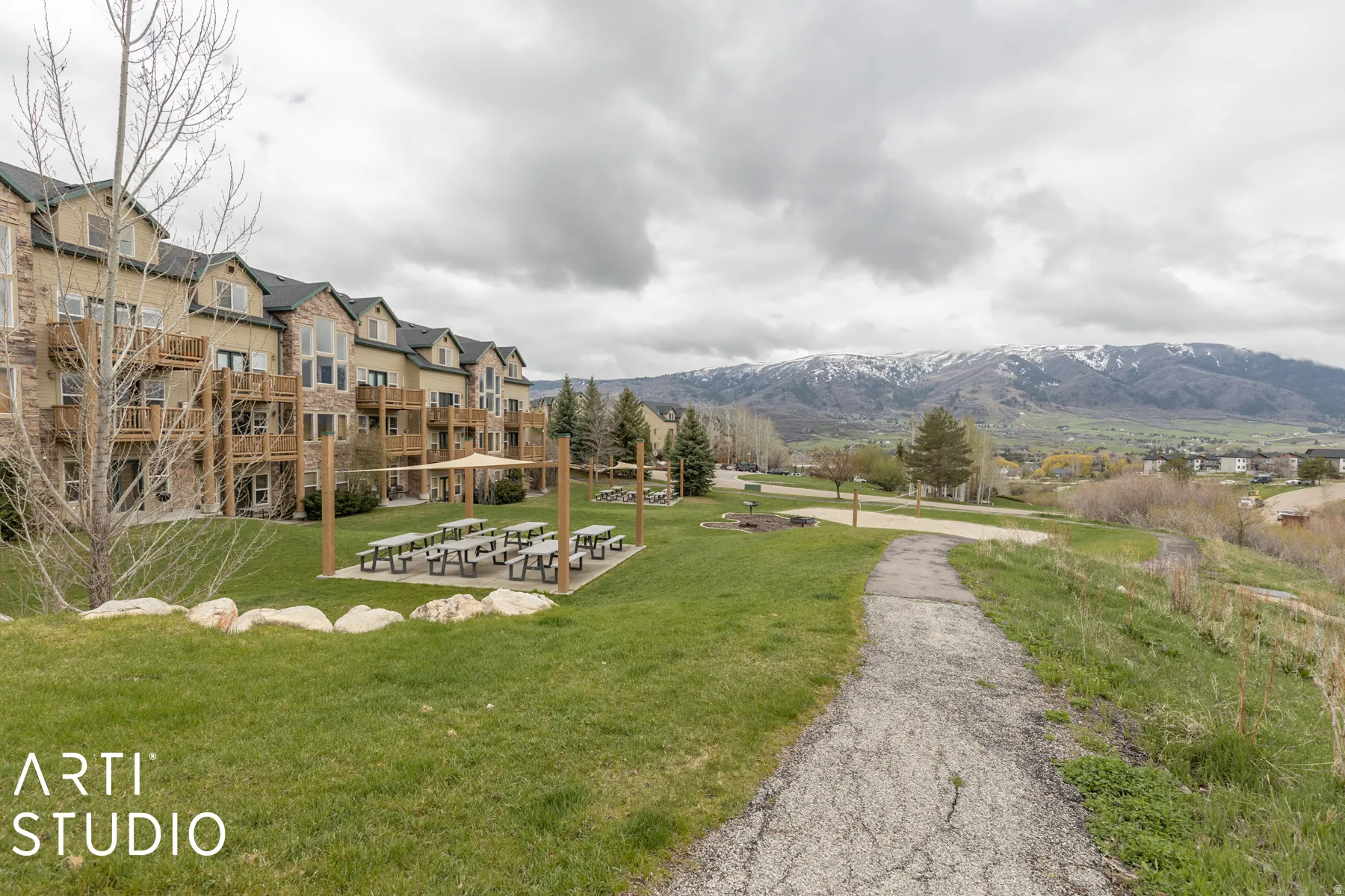 Multi-story building with stone and siding exterior, private balconies, an outdoor dining area with picnic tables and shade structures, and a paved pathway