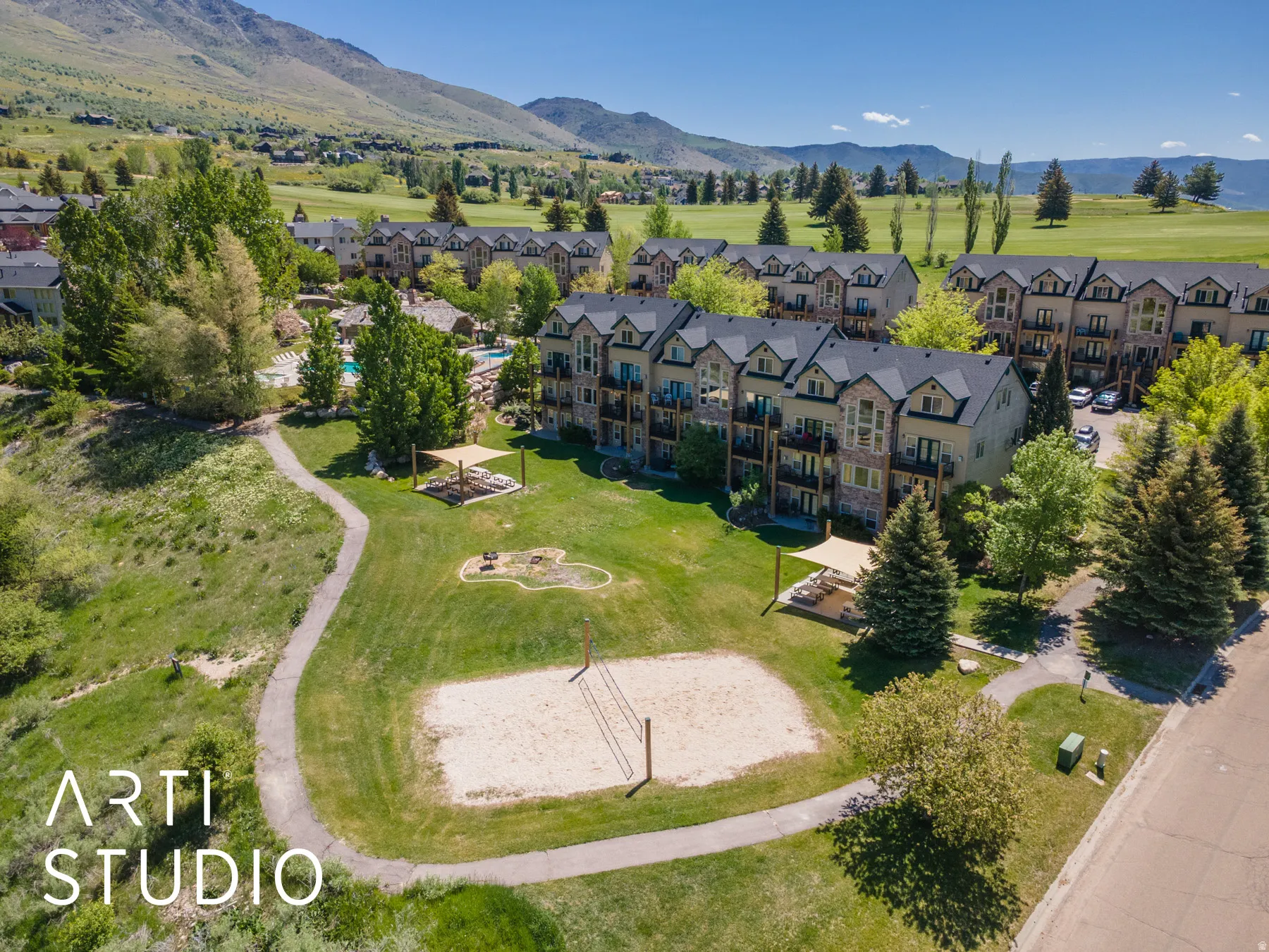 Expansive green space featuring a sand volleyball court, paved walking paths, and shaded patio areas