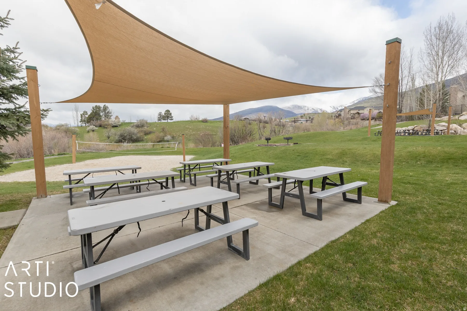 Outdoor recreational space featuring multiple picnic tables, a shade sail structure, and an adjacent sand volleyball court
