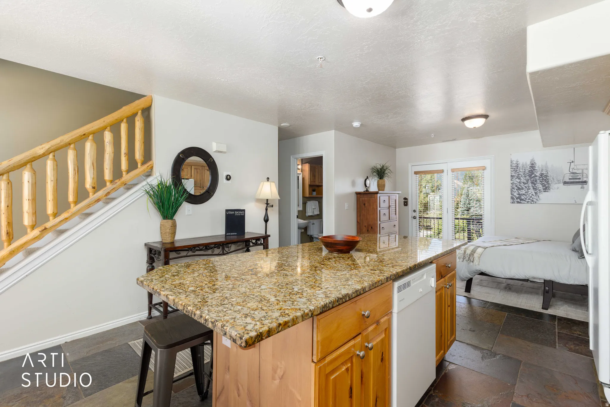 Kitchen island with stone countertop and wood-finish cabinetry, integrated dishwasher, slate tile flooring, and a wood-finish staircase with rustic balusters