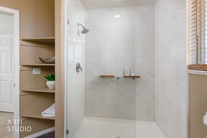 Newly renovated glass-enclosed shower with white tiled walls, a rain showerhead, and integrated shelving
