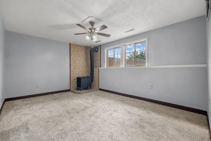 Carpeted room featuring a wood-burning stove with brick surround, dual windows, and a ceiling fan