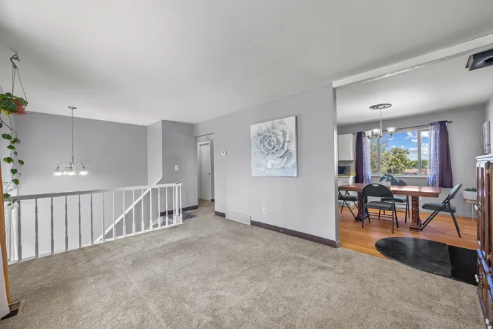 Carpeted living area featuring a white railing, a contemporary chandelier, and a framed opening to an adjacent room with wood-finish flooring and a window