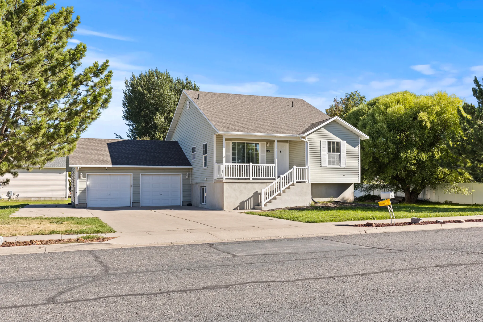 Light-colored siding exterior, two-car garage, white railing porch, mature landscaping, and a concrete driveway