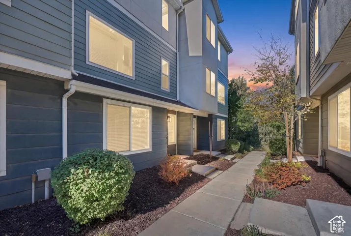 Multi-story exterior featuring blue horizontal siding, white trim, multiple windows, and a concrete walkway bordered by landscaped garden beds