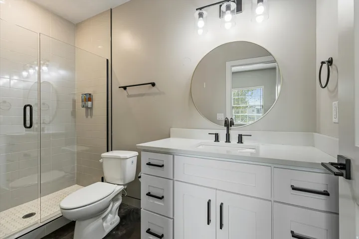 Bathroom featuring a glass-enclosed shower, white vanity with integrated sink, stone countertop, matte black fixtures, and a circular mirror