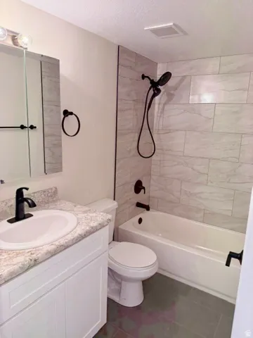 Bathroom featuring a single-sink vanity with a speckled countertop, a white toilet, a bathtub with a tiled surround, and matte black fixtures