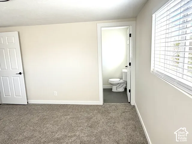 Carpeted room with a window featuring blinds and a white door, with an adjacent bathroom featuring a toilet and dark tile flooring