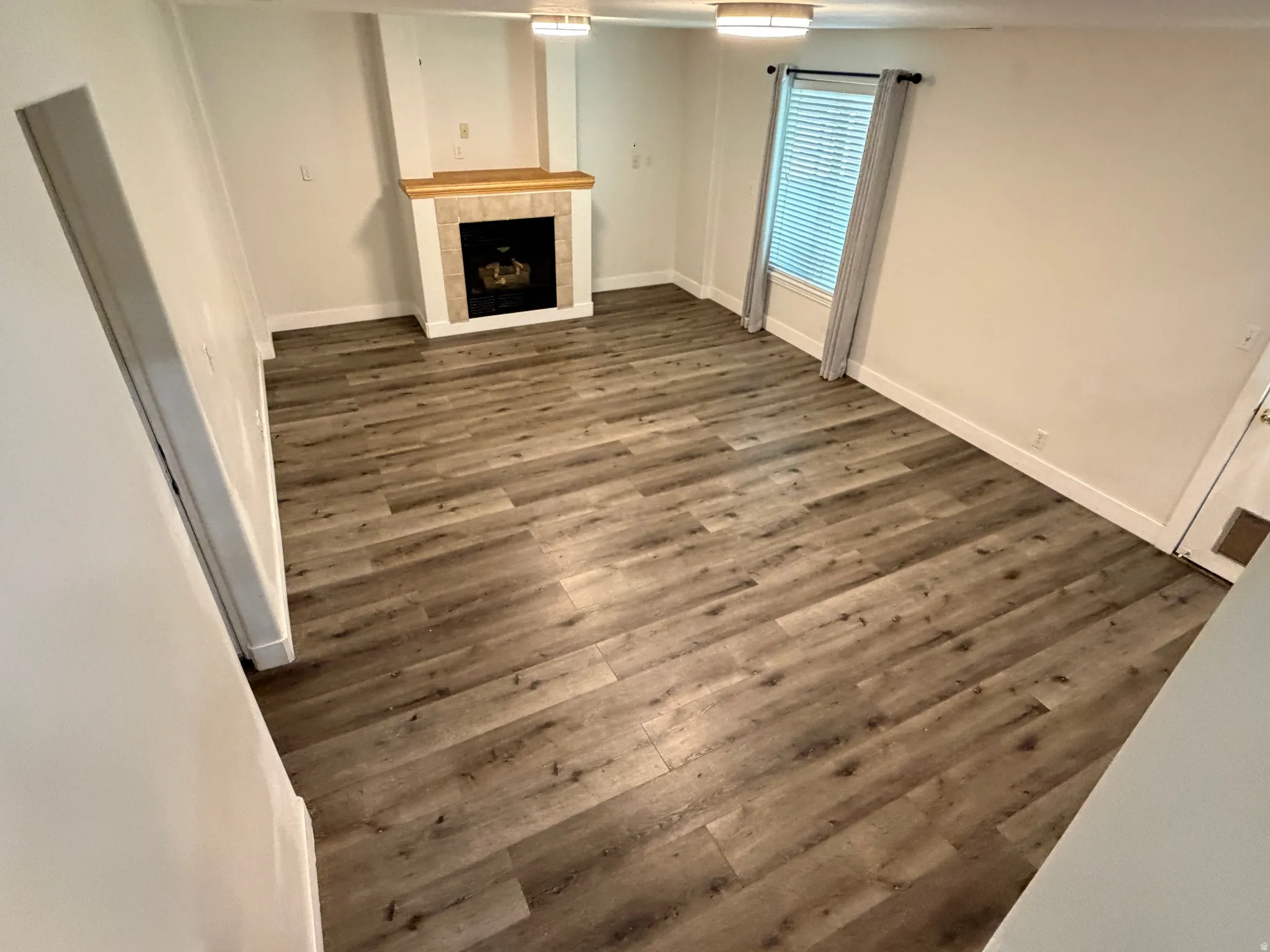 Wood-finish flooring, fireplace with tile surround and wood mantel, white baseboards, window with blinds, and flush mount lighting