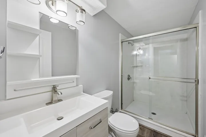 Full bathroom featuring a single vanity with integrated sink, wood-finish cabinetry, a framed mirror with integrated shelving, and a glass-enclosed shower with built-in shelving