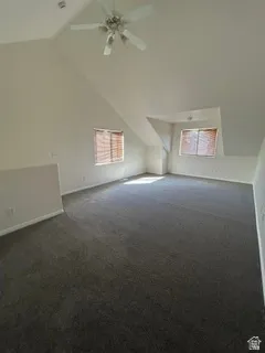 Spacious room featuring vaulted ceilings, ceiling fan, two windows with blinds, and dark carpet flooring