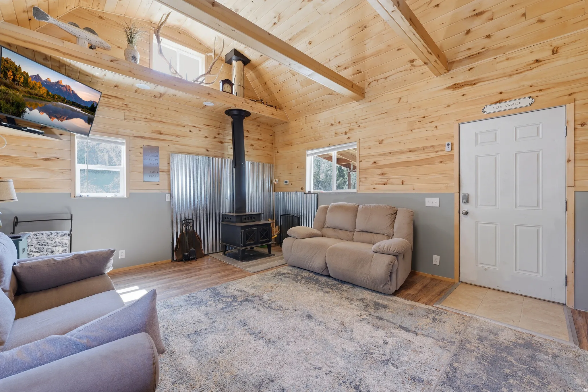 Vaulted wood-paneled ceilings with exposed beams, wood-finish flooring, and a black wood-burning stove with a corrugated metal surround
