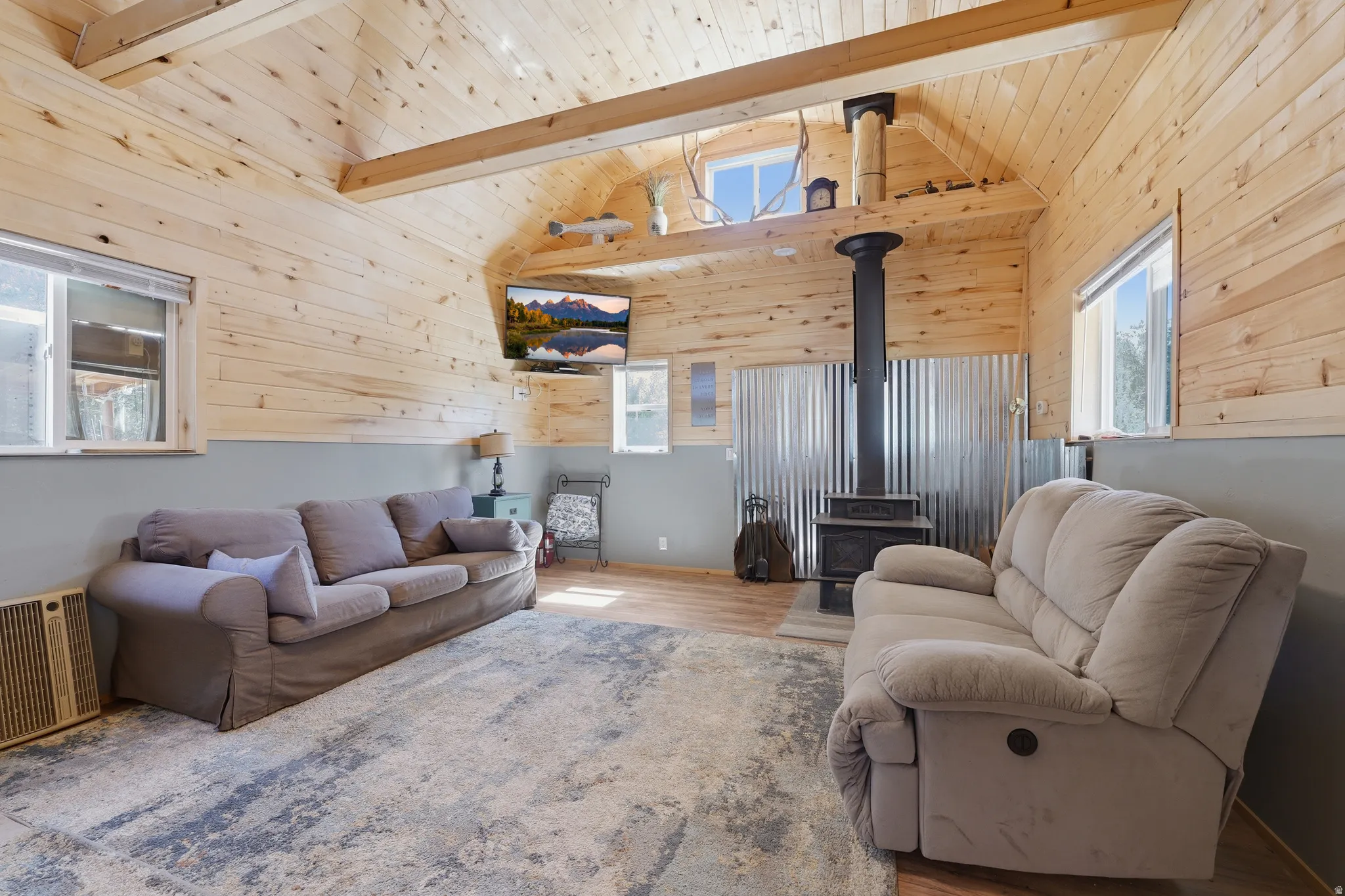 Vaulted wood plank ceiling, exposed structural beams, wood-finish flooring, and a wood-burning stove with corrugated metal backing
