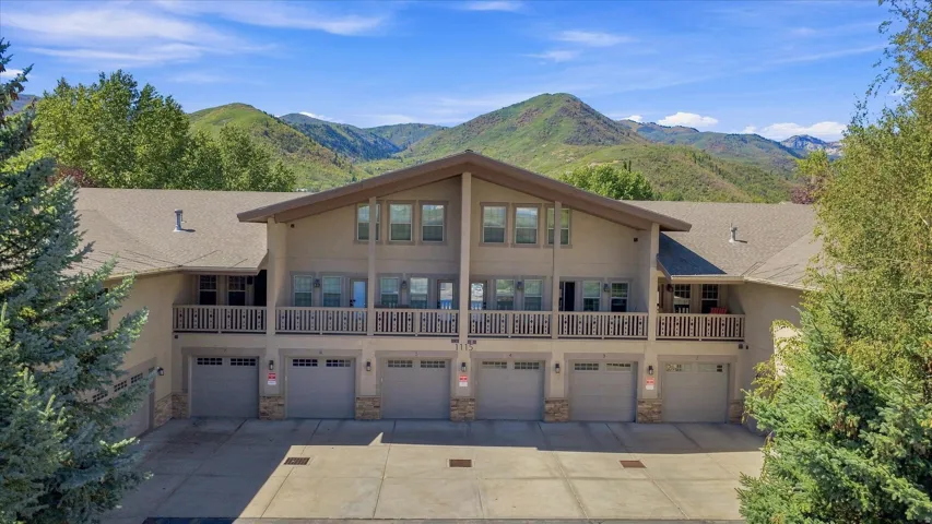Multi-level residential building featuring a neutral-toned exterior, multiple attached garages, upper-level balconies with railings, prominent windows, and a backdrop of verdant mountains