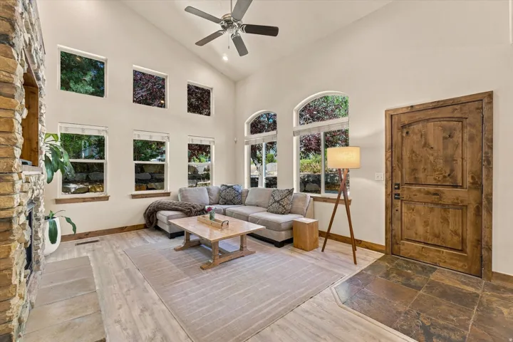 Vaulted ceiling with a ceiling fan, stone-clad fireplace with hearth, wood-finish flooring, multiple windows, and a wood entry door