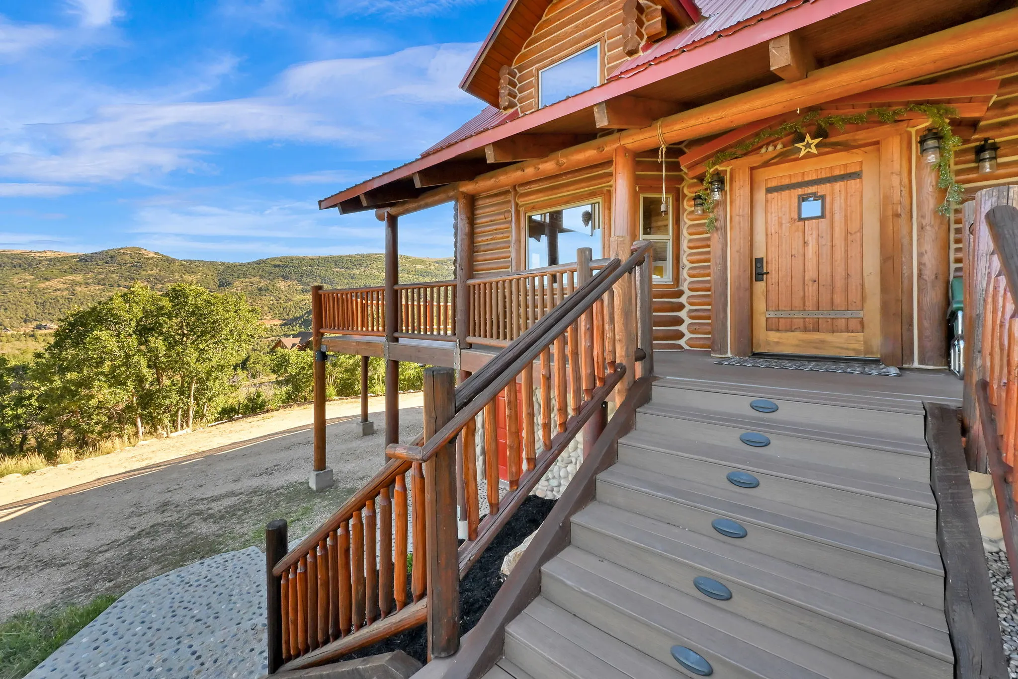 Log cabin exterior with a visible mountain backdrop, featuring a covered porch, wood plank front door, and exterior stairs with integrated lighting