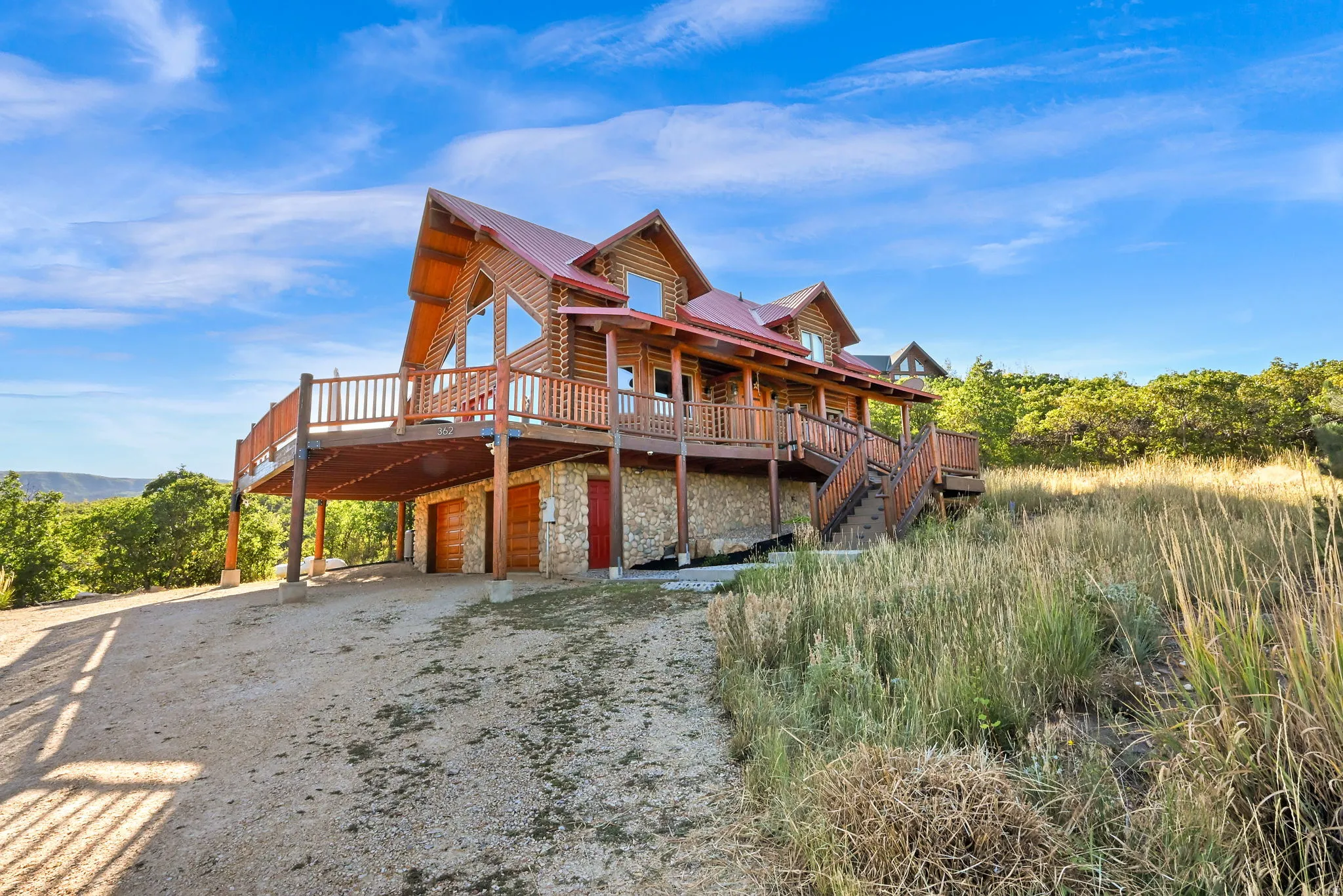 Log home exterior with a red metal roof, expansive wraparound deck with wooden railings, and a stone foundation