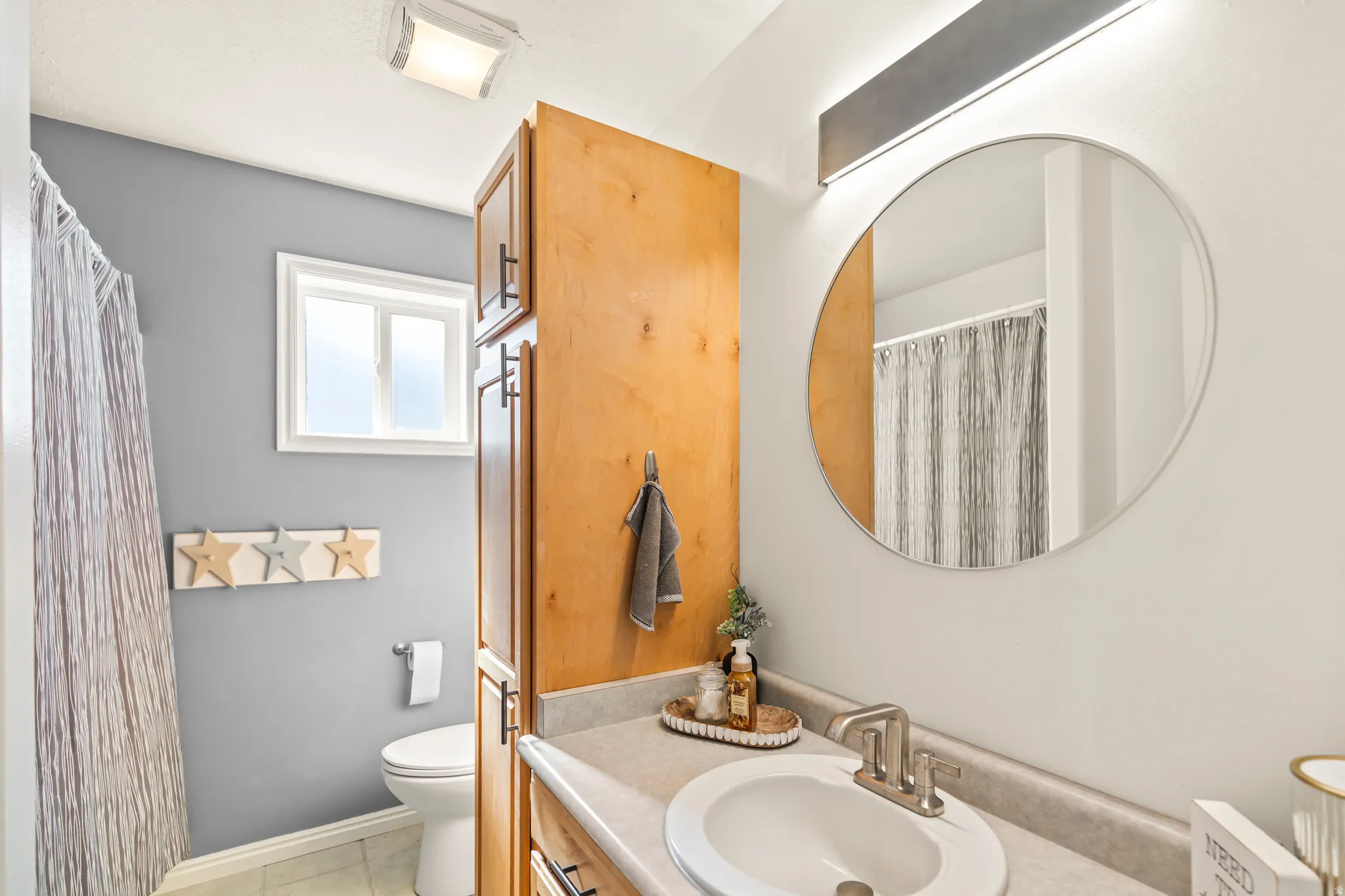 Light wood-finish cabinetry, built-in storage, white framed window, round vanity mirror, and an overhead light bar