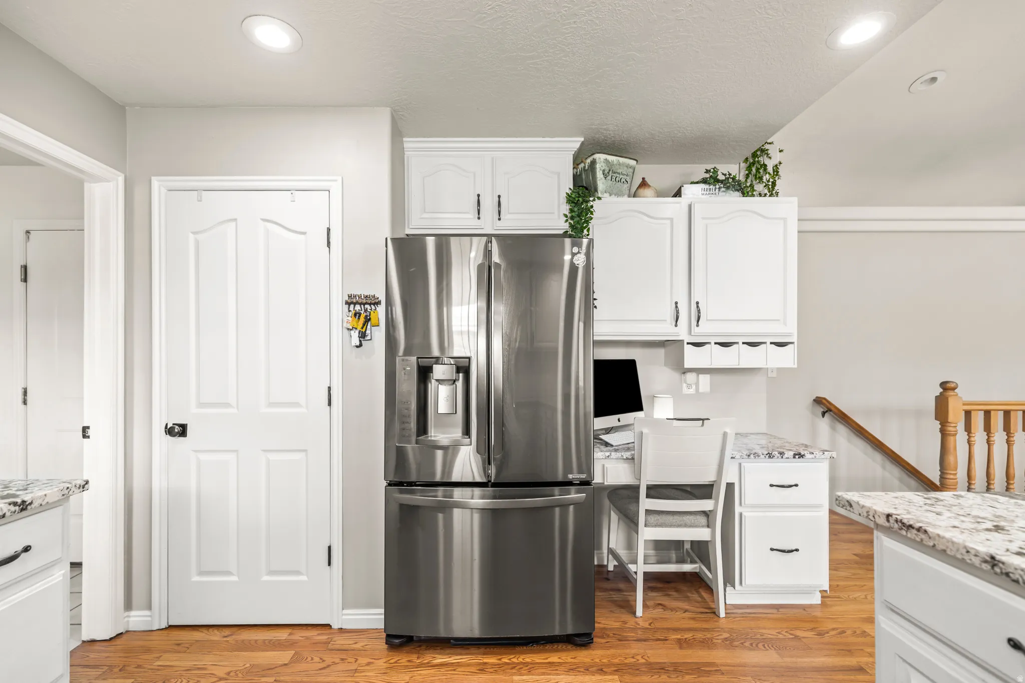 Stainless steel French door refrigerator, white cabinetry, built-in desk area with drawers, recessed lighting, and wood-finish flooring