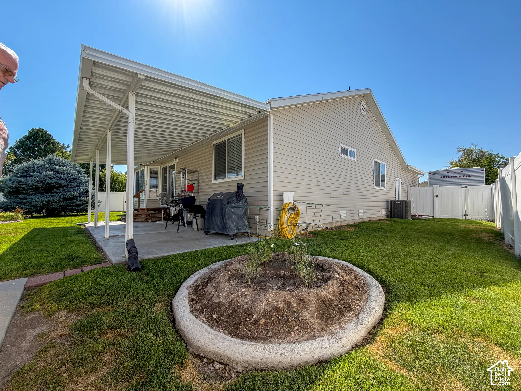 Covered concrete patio, vinyl siding, mature shrubbery, partial white fencing, and an established lawn