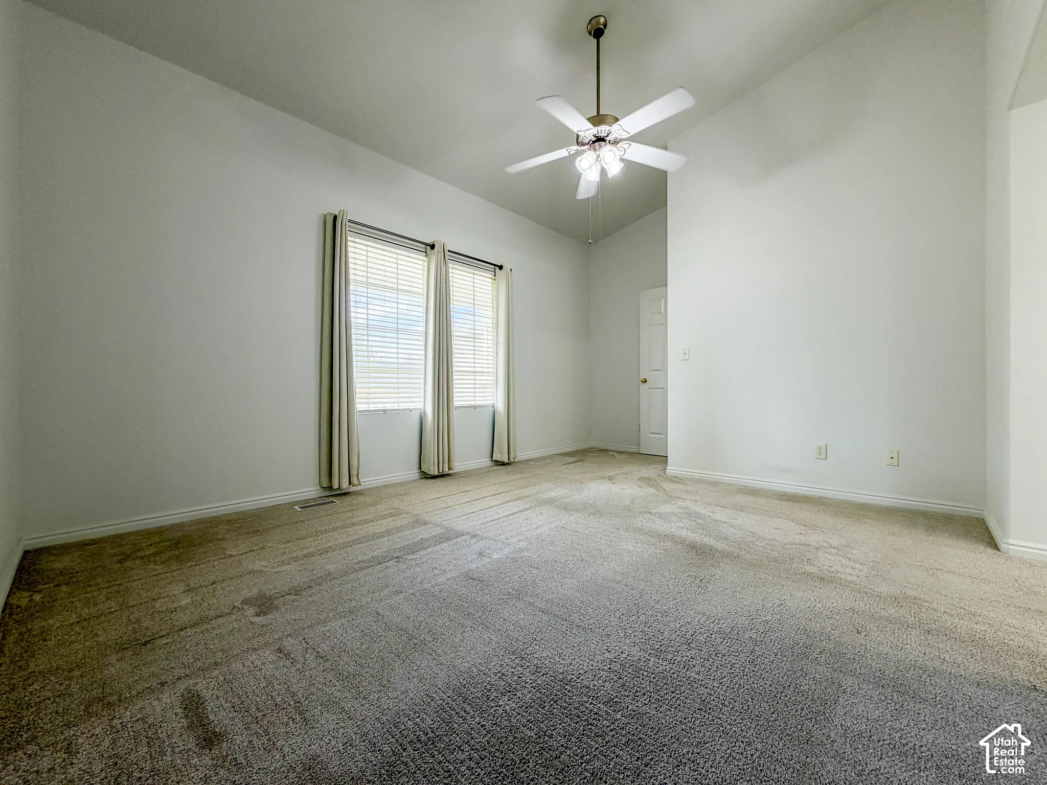 Vaulted ceiling with a ceiling fan light fixture, dual windows with blinds, and neutral tone carpeting