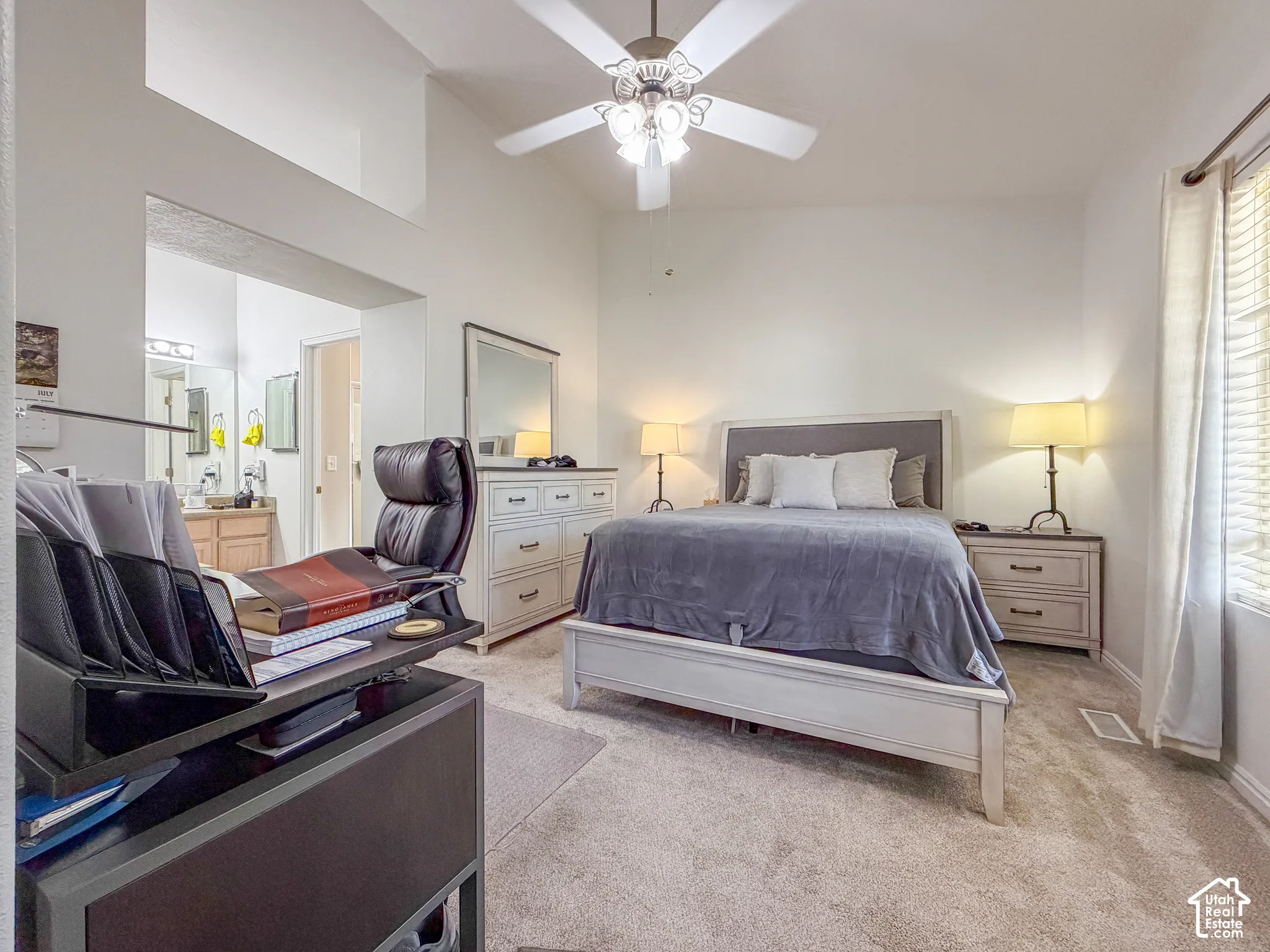 Vaulted ceiling with ceiling fan, plush carpet flooring, window with blinds, and built-in vanity with dual sinks visible in the background