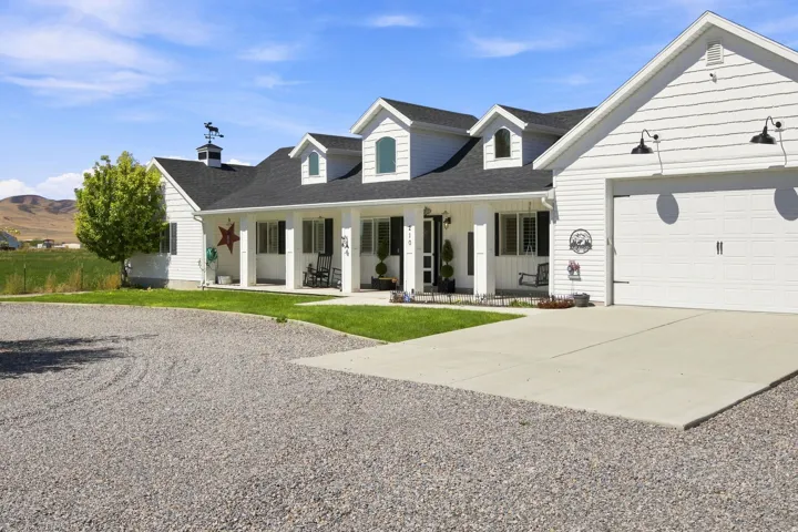 White siding exterior with a dark shingle roof, covered front porch, attached garage with overhead lighting, and a gravel driveway
