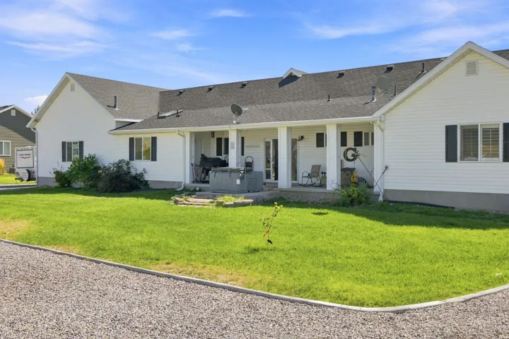 White siding exterior with dark shutters, covered patio, gravel driveway, and a spacious lawn