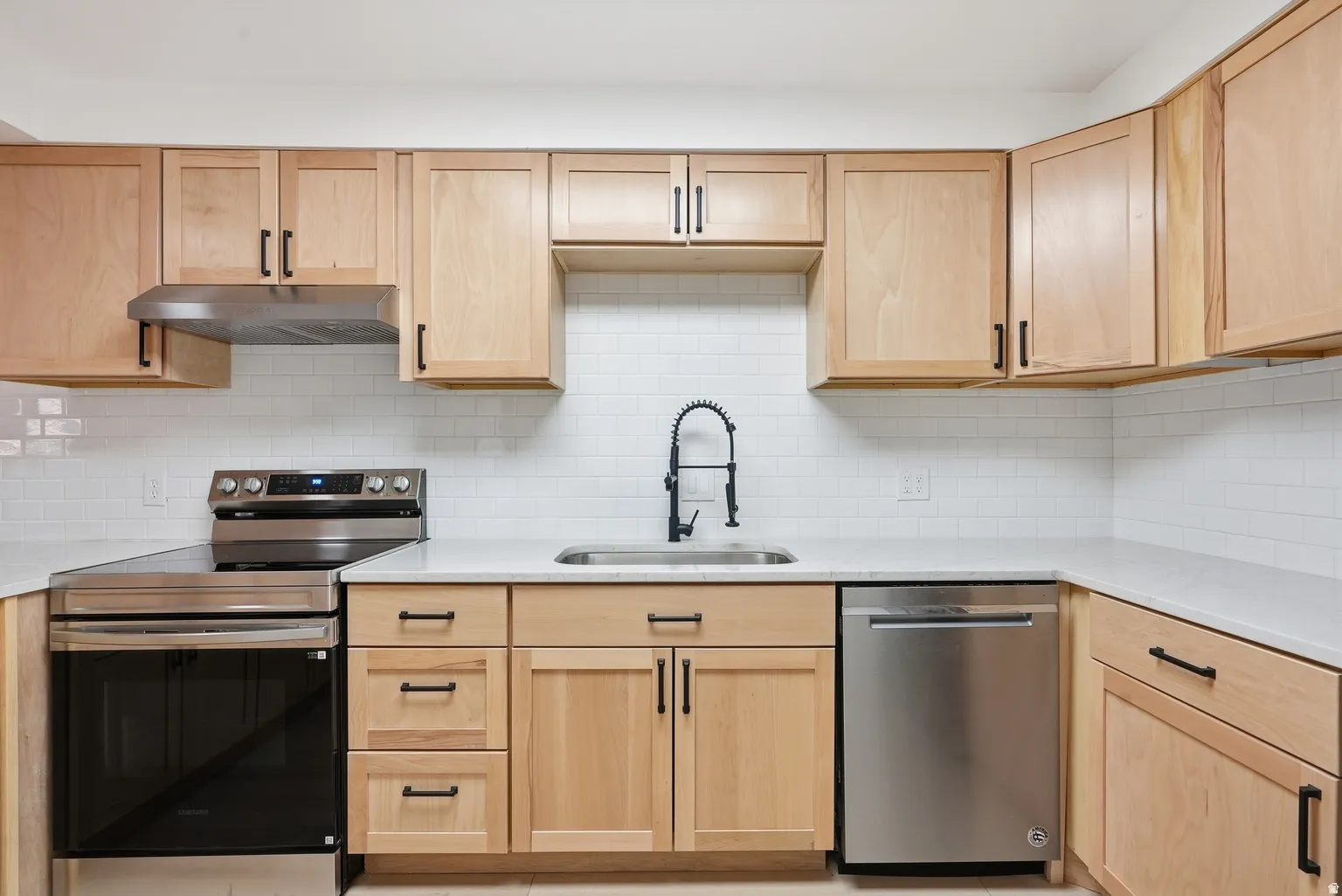 Light wood-finish cabinetry with matte black hardware, stainless steel appliances, white subway tile backsplash, and light-toned countertops