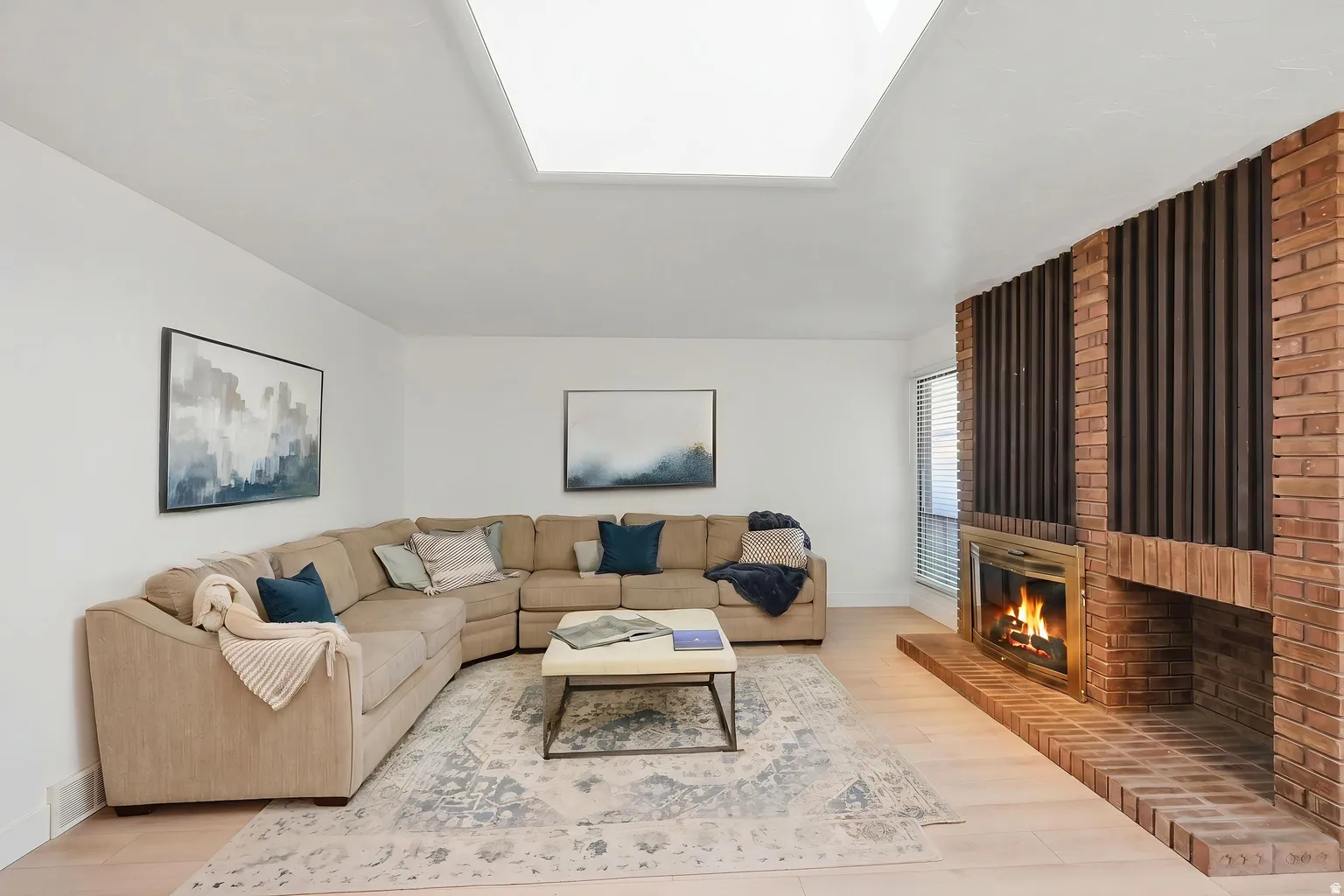Recessed skylight, light wood-finish flooring, brick fireplace with a wood mantel, and integrated window blinds