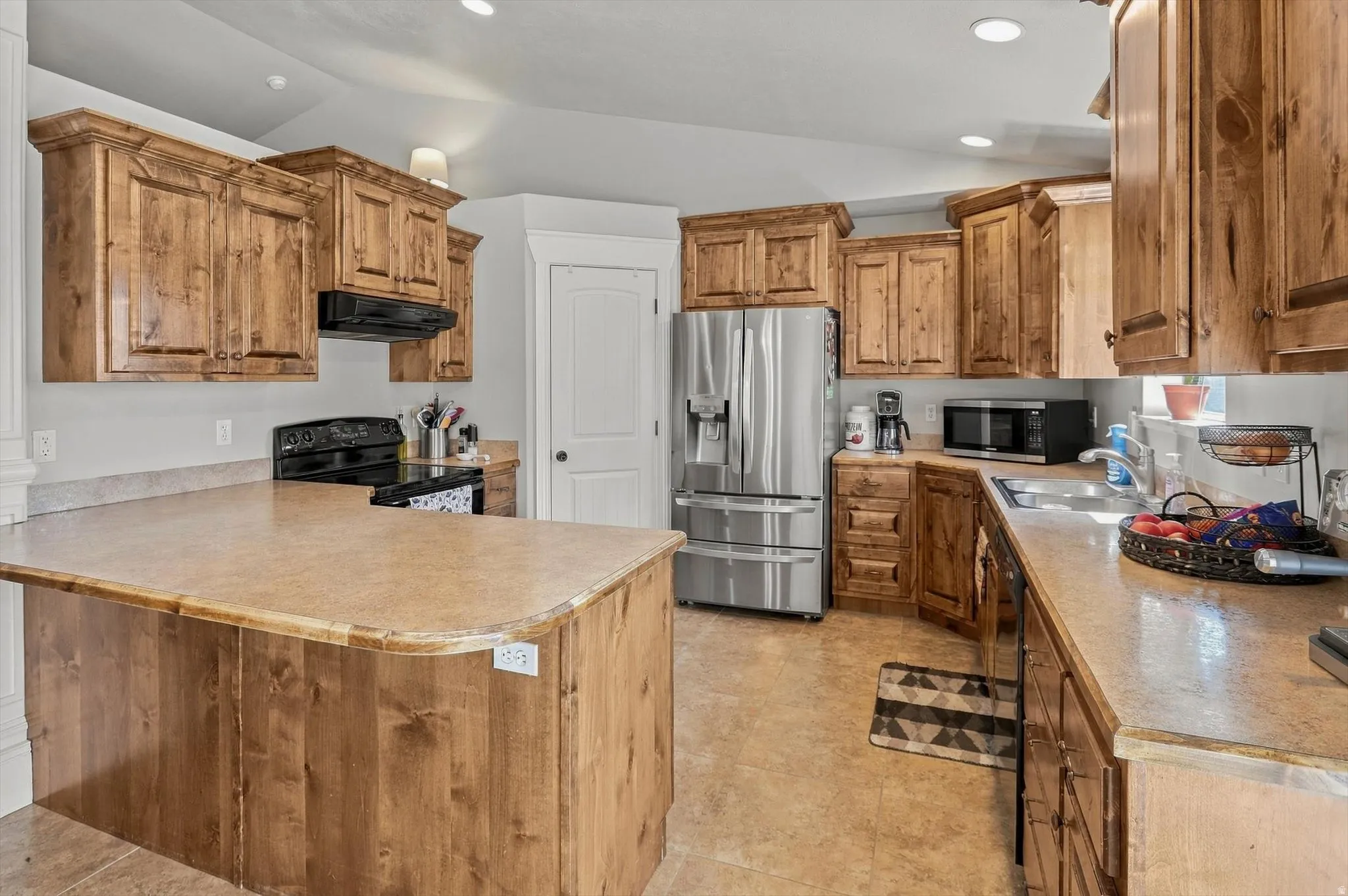 Wood cabinetry and a matching island with a bar-height counter, stainless steel refrigerator, dark cooktop, and recessed lighting