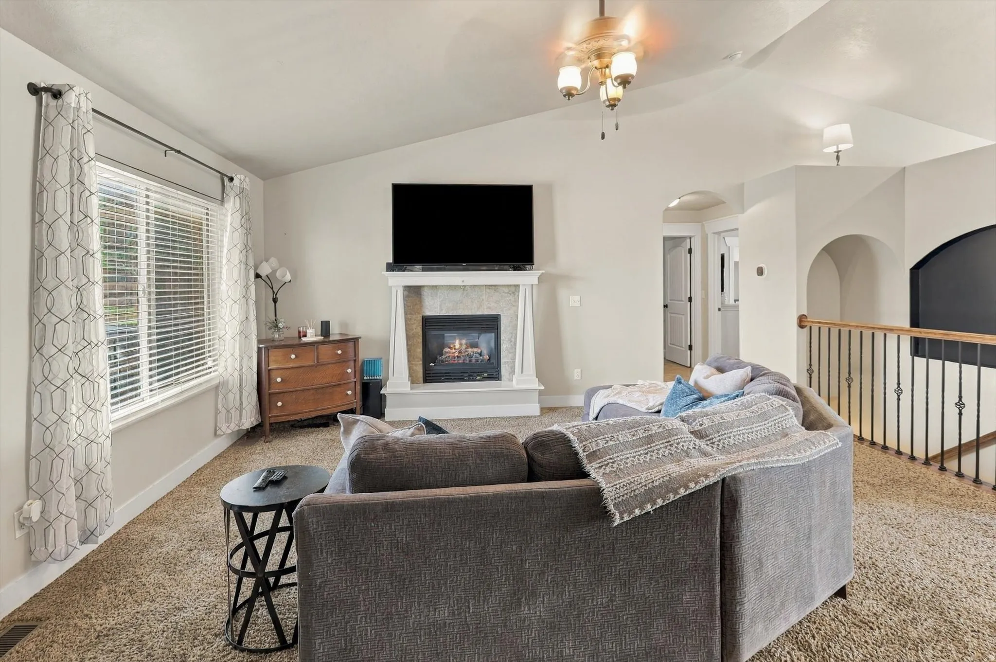 Vaulted ceiling with a chandelier, fireplace with a mantle and tile surround, large window with blinds, carpeted flooring, and an arched doorway
