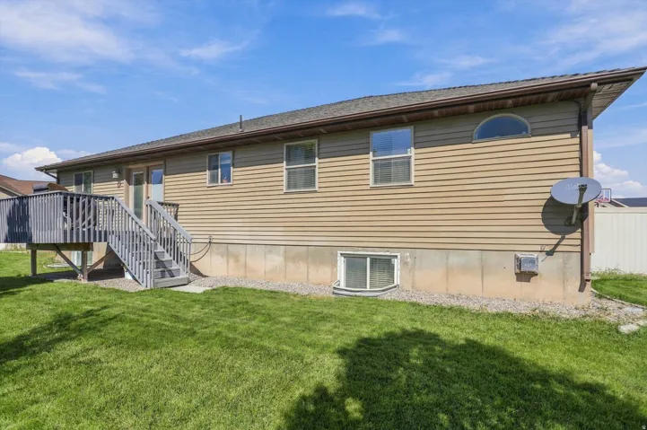 Expansive backyard featuring a manicured lawn, a raised wooden deck with stairs, tan horizontal siding, and a basement window