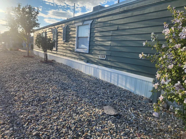 Siding exterior with light-colored skirting, gravel landscaping, and mature trees
