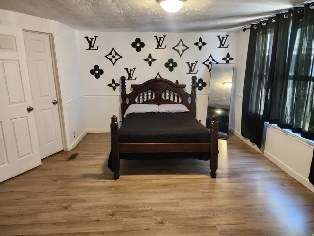 Spacious bedroom featuring refinished wood flooring, a decorative accent wall with geometric patterns, and abundant natural light from large windows dressed in contemporary black drapery
