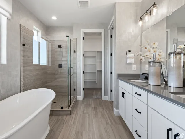 Freestanding soaking tub, glass-enclosed shower with tiled walls, dual vanity with solid surface countertop, white cabinetry with black hardware, and wood-finish flooring