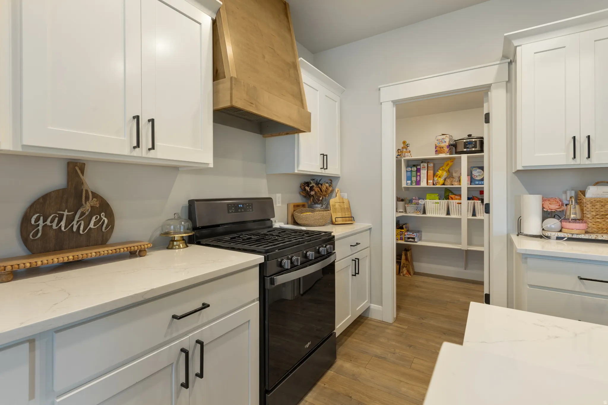 White cabinetry with matte black hardware, light-toned countertops, wood-finish flooring, a built-in gas range, and a walk-in pantry with shelving
