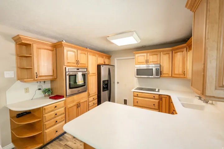 Light wood cabinetry with crown molding, integrated stainless steel appliances, white solid surface countertops, a double basin sink, and wood-finish flooring