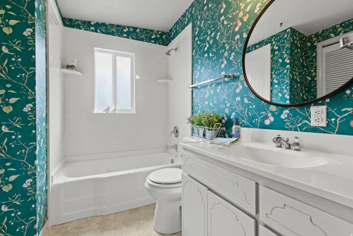 Bathroom featuring a built-in tub and shower combination, a window with frosted glass, a vanity with a white countertop and integrated sink, a circular wall-mounted mirror, and patterned wallpaper