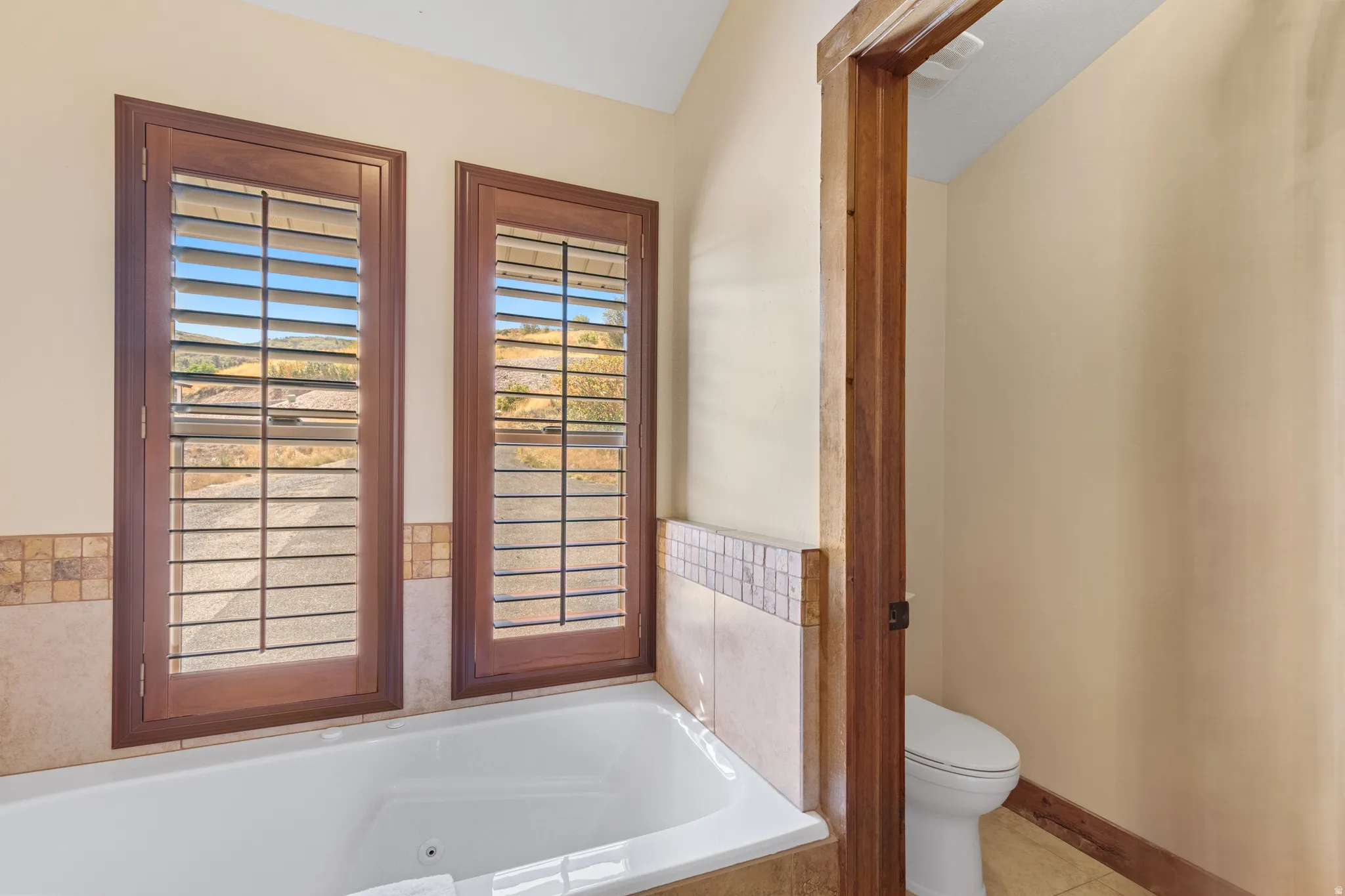 Built-in soaking tub with a tiled surround featuring a mosaic accent, two windows with wooden plantation shutters, and a private water closet with a toilet