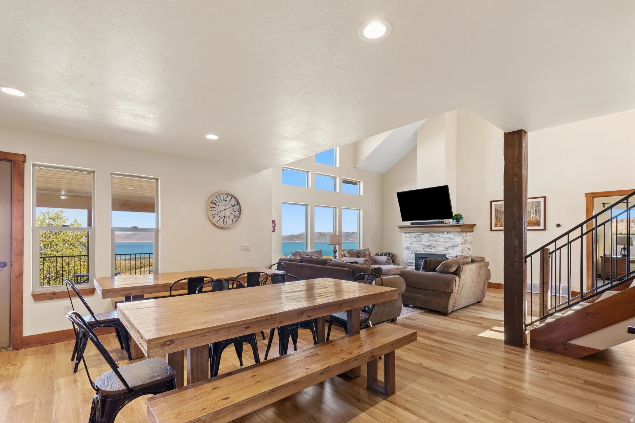 Vaulted ceiling with recessed lighting, large windows, wood-finish flooring, stone-clad fireplace, and an open staircase with a wrought iron railing