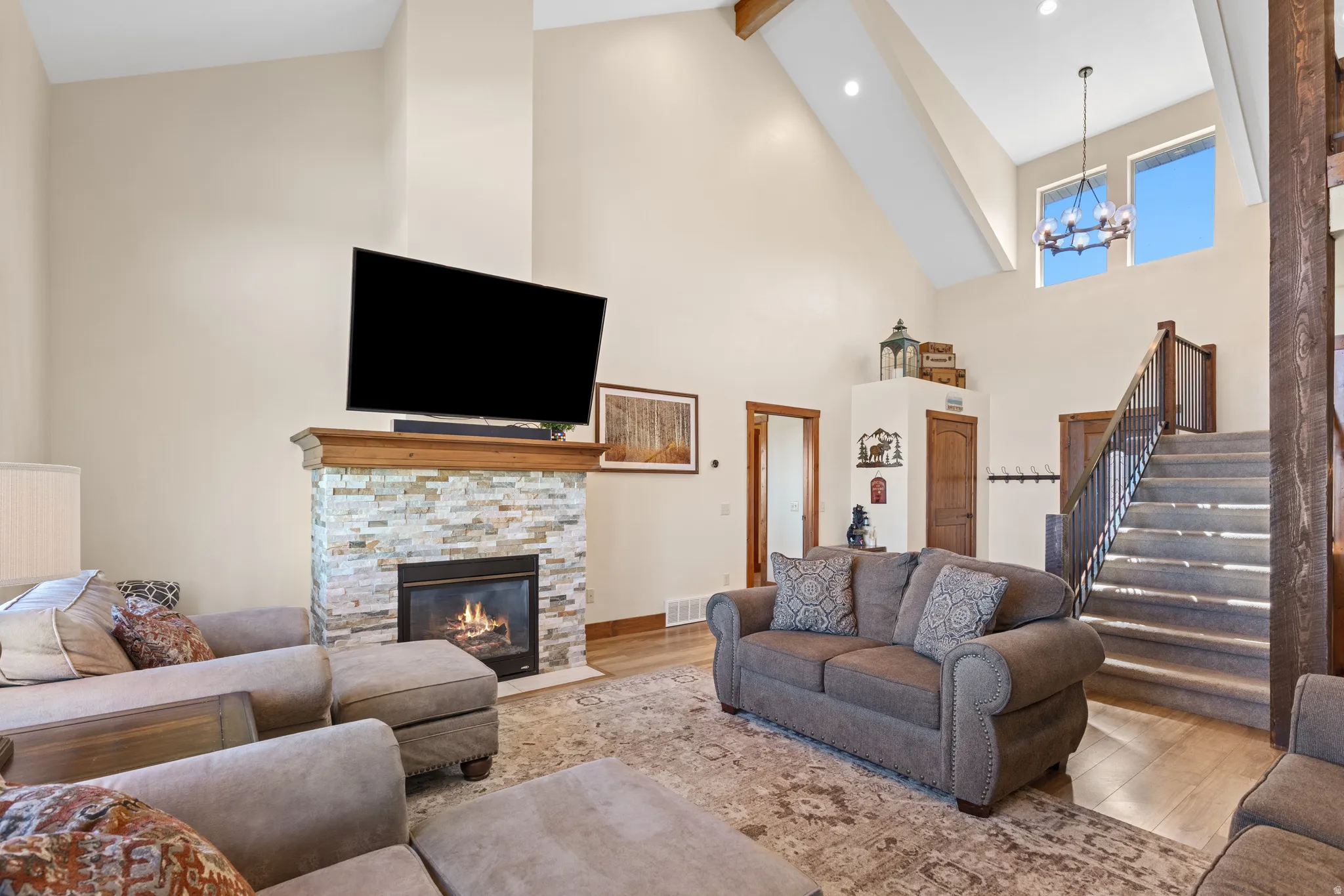 Vaulted ceiling with exposed beams, stacked stone fireplace with wood mantel, wood-finish flooring, two-story windows, and a chandelier