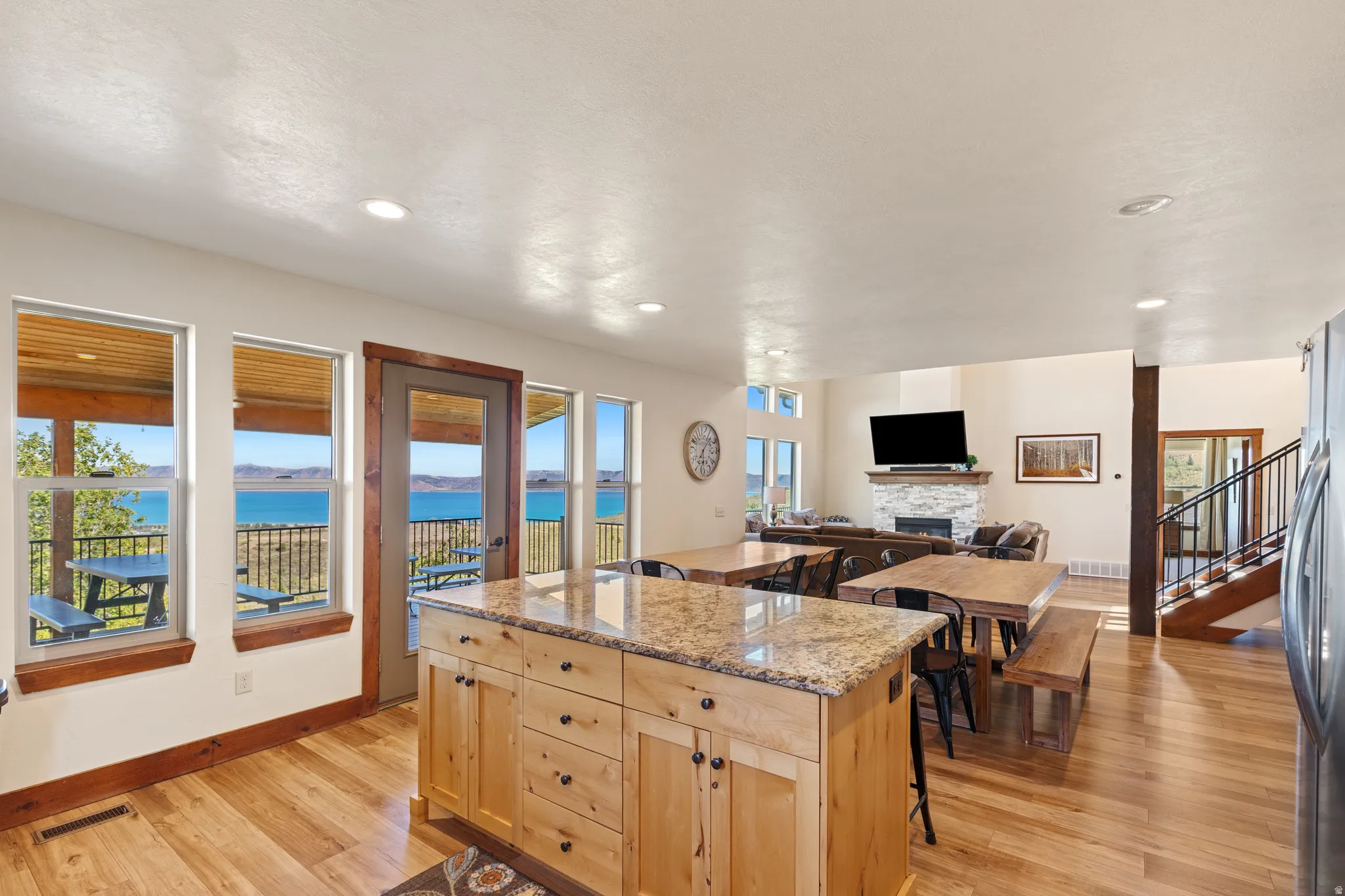 Gourmet kitchen island with granite countertop, wood-finish flooring, multiple windows providing a water backdrop, stone-wrapped fireplace, and a prominent staircase with metal railings