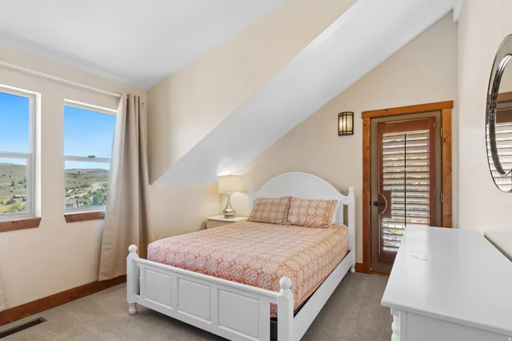 Sloped ceiling room featuring dual windows, wood-trimmed door with louvered shutters, wall-mounted sconce, and carpeted flooring