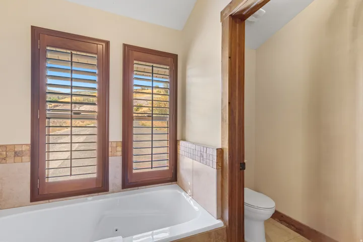 Built-in soaking tub with a tiled surround featuring a mosaic accent, two windows with wooden plantation shutters, and a private water closet with a toilet