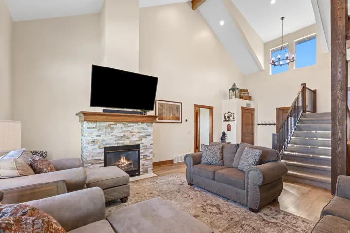 Vaulted ceiling with exposed beams, stacked stone fireplace with wood mantel, wood-finish flooring, two-story windows, and a chandelier