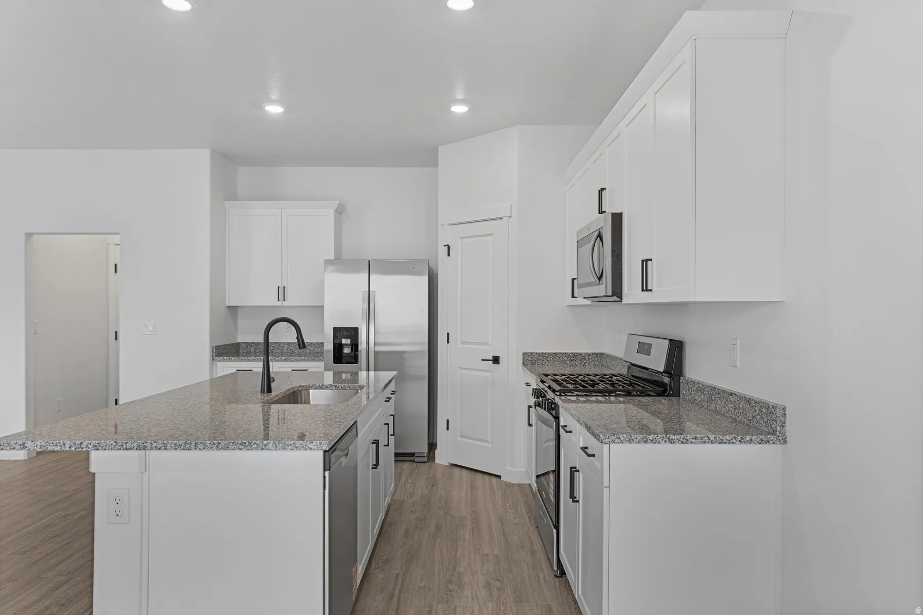 Kitchen featuring white cabinetry, granite countertops, stainless steel appliances, a kitchen island with an undermount sink, and wood-finish flooring
