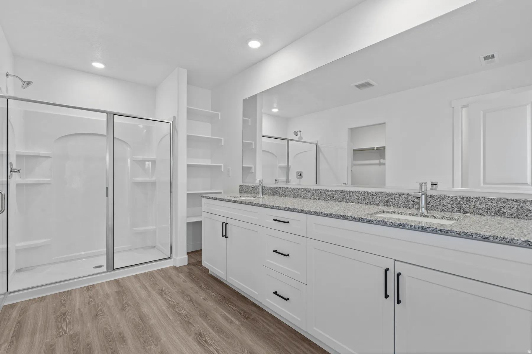 Dual vanity with granite countertop, wood-finish flooring, glass-enclosed shower, built-in shelving, and recessed lighting