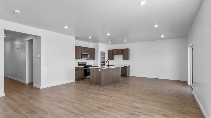 Open-concept living area with wood-finish flooring, recessed lighting, and a kitchen featuring dark wood cabinetry, a center island with a sink, and stainless steel appliances
