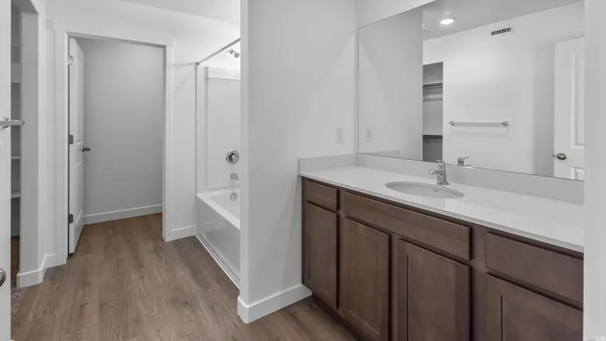 White acrylic tub/shower combination, wood-finish flooring, dark wood-finish vanity with an undermount sink, light-colored countertop, and a large wall-mounted mirror